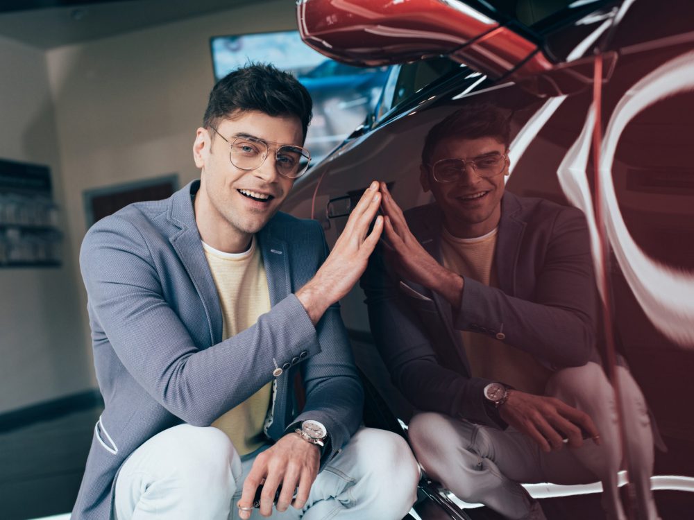 happy stylish man in glasses sitting near red automobile